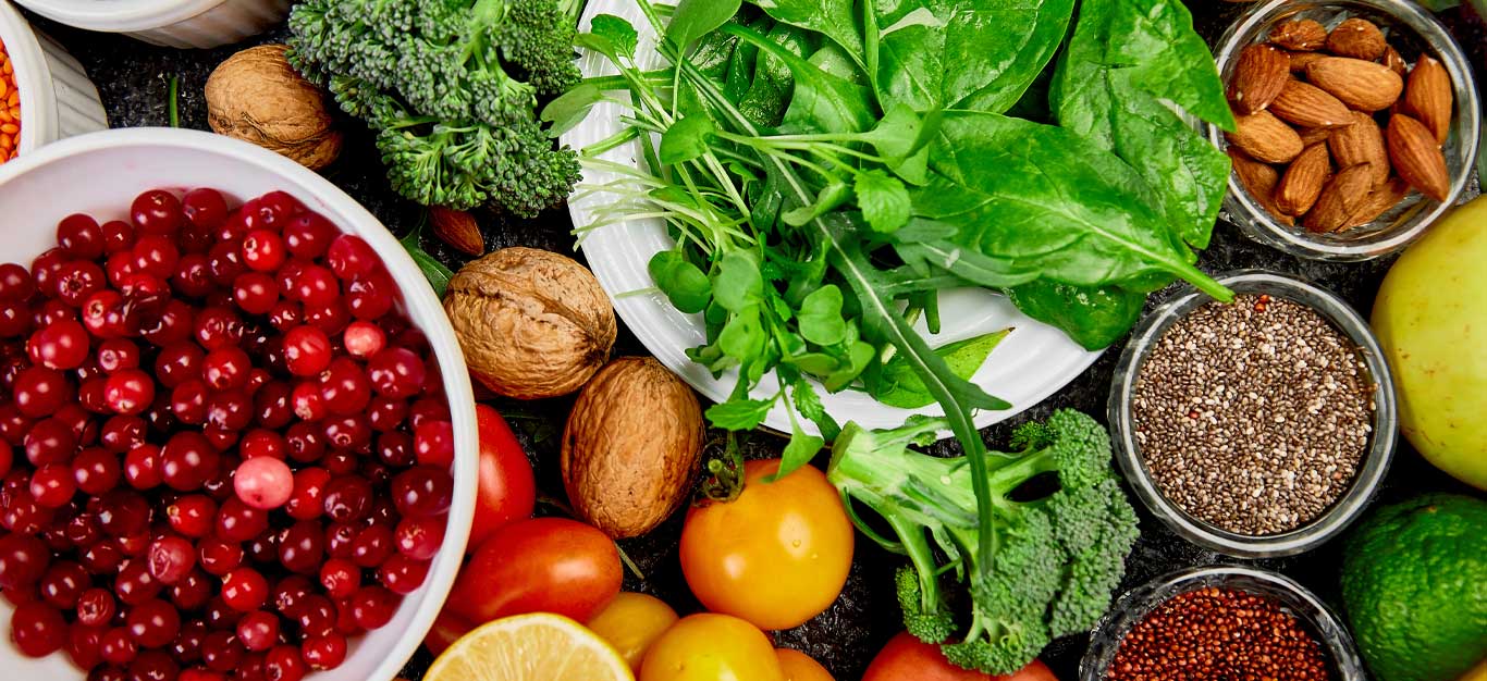 A bowl of cranberries surrounded by fresh tomatoes, broccoli, spinach, walnuts, almonds, and small bowls of chia seeds and quinoa