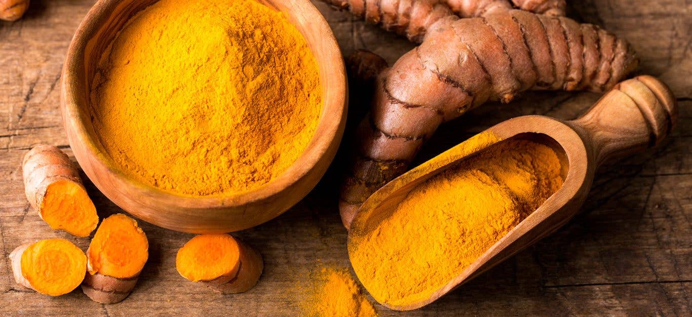 A table with an array of turmeric on it, including a wooden bowl of dried ground turmeric, with a scoop next to it filled with more dried turmeric, and a knob of fresh turmeric root and some fresh turmeric slices