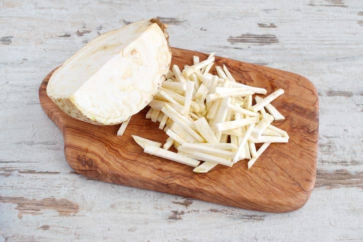 Root celery cut into small pieces with the half of the whole celery root on a olive wood cutting board on a light wooden background. 