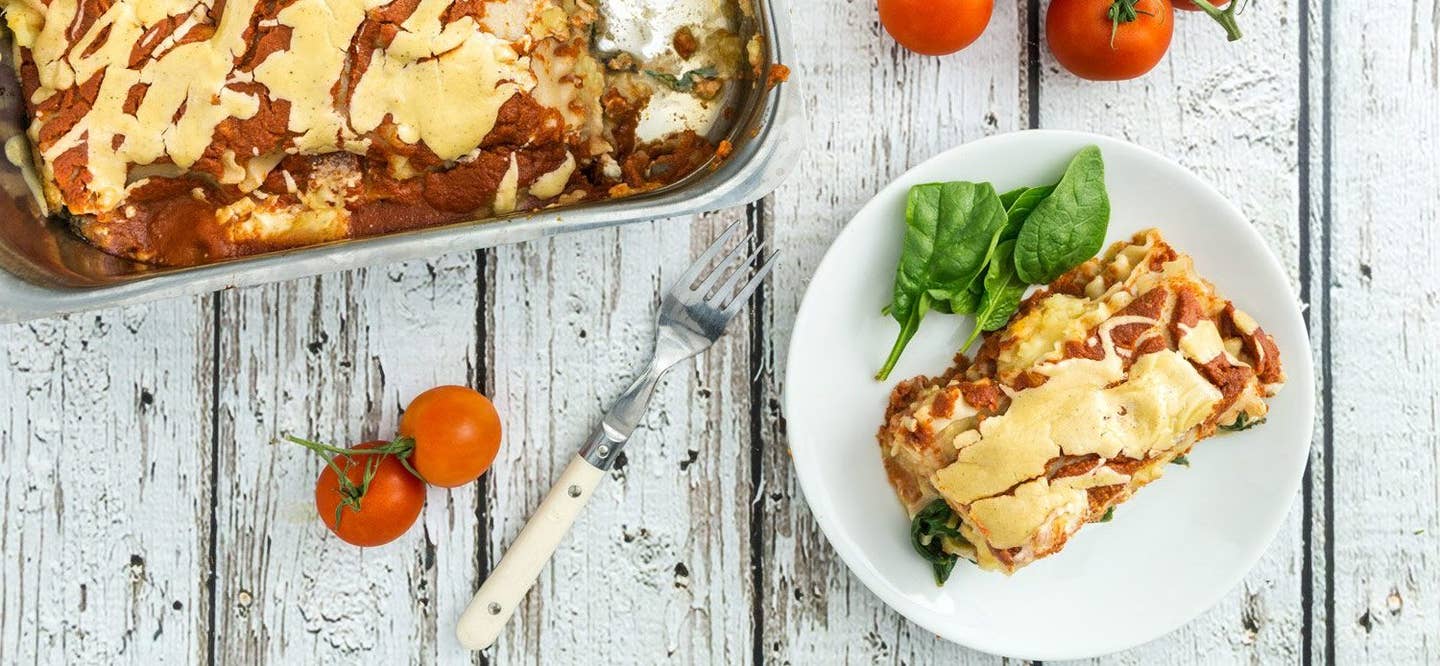 Sweet Potato Lasagna slice on a white plate next to a large tray of lasagna on a white wooden tabletop