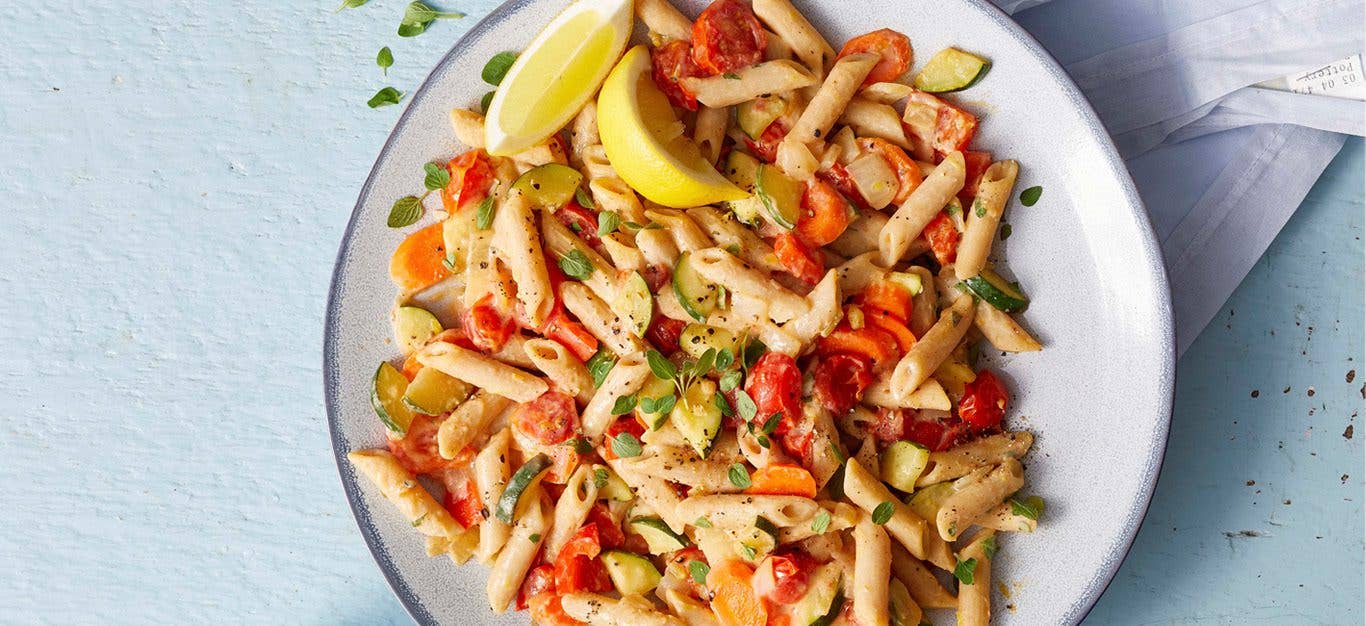 Lemon Hummus Pasta: Penne pasta tossed with a creamy hummus-based dressing, tossed with chopped red bell pepper. Served on a white plate, with lemon wedges on the side.