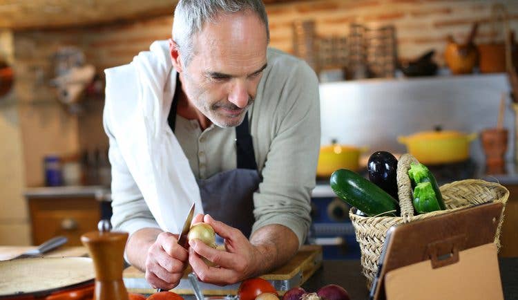 A middle-aged man leans over his kitchen counter reading a recipe off an iPad while peeling an onion