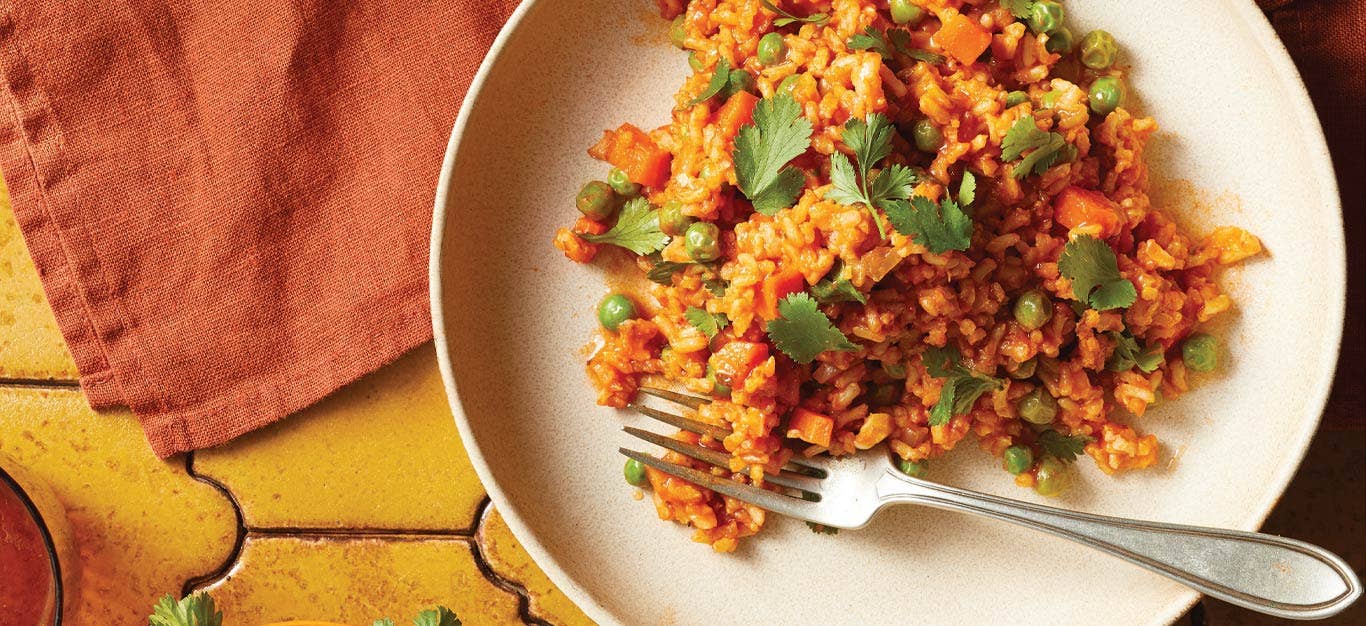 Mexican-Style Brown Rice on a while plate with a metal fork sitting on a yellow tile tabletop