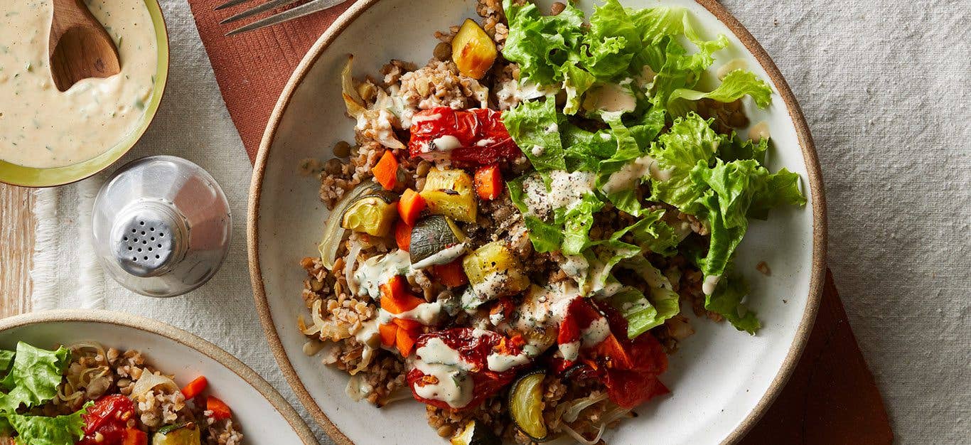 vegan mujadara bowls with chopped lettuce, colorful vegetable medley, lentils, and a creamy homemade tahini sauce with parsley - shown in a white bowl in sunlight