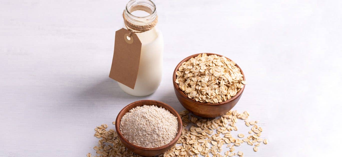 A clean white surface with a carafe of oat milk, a small bowl of oat flour, and a slightly larger bowl of whole oats