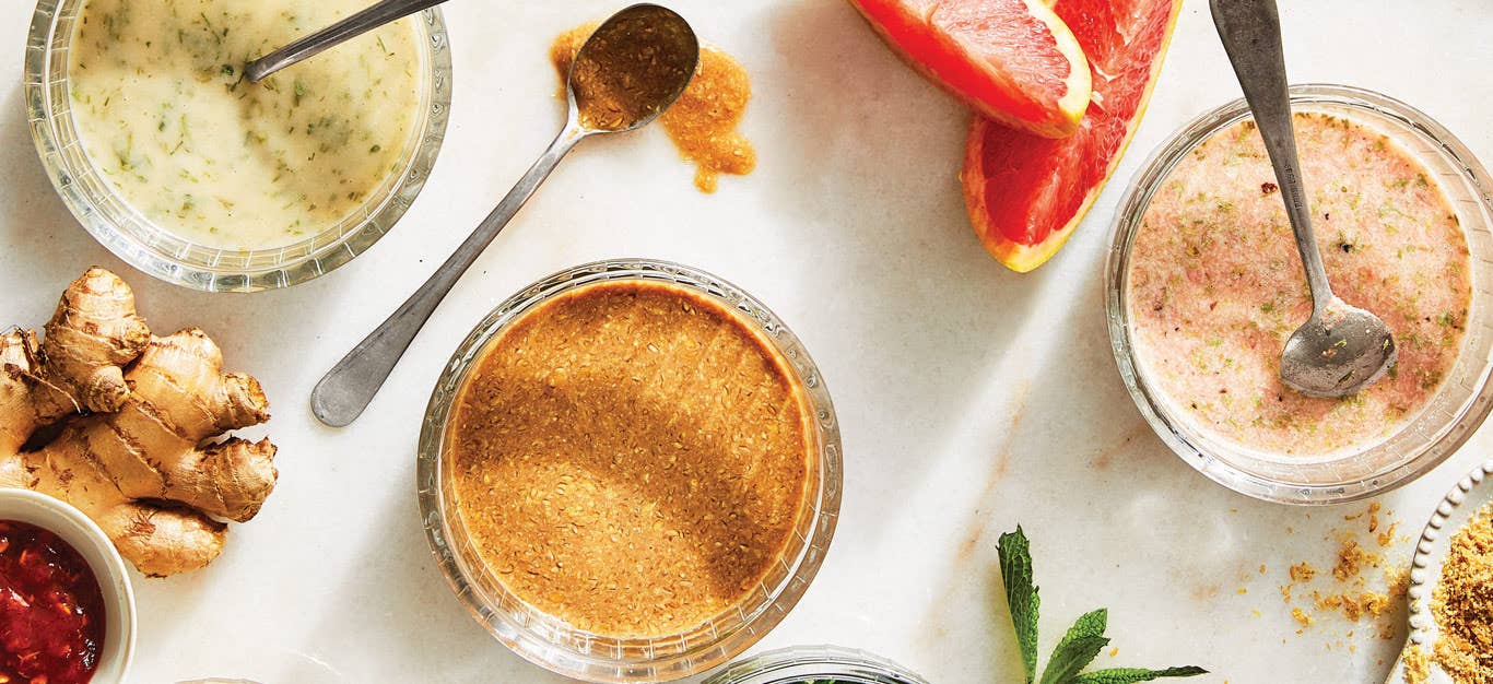 An array of homemade salad dressings made without oil, including vegan ranch, balsamic vinaigrette, and a grapefruit tarragon, in small glass bowls with spoons in them and some of the ingredients shown beside them