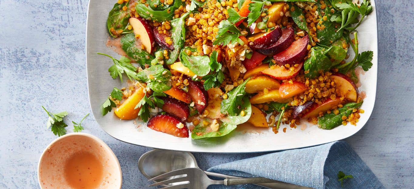 Plum-Spinach Salad with Curried Lentils on a white plate with utensils and a small bowl of dressing