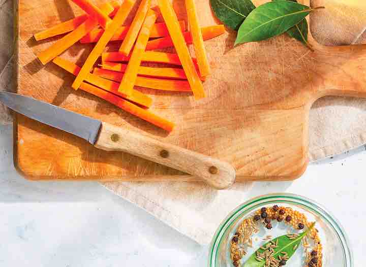 cutting board with trimmed carrots, bay leaves, whole black peppercorns, and cumin seeds, preparing for pickling