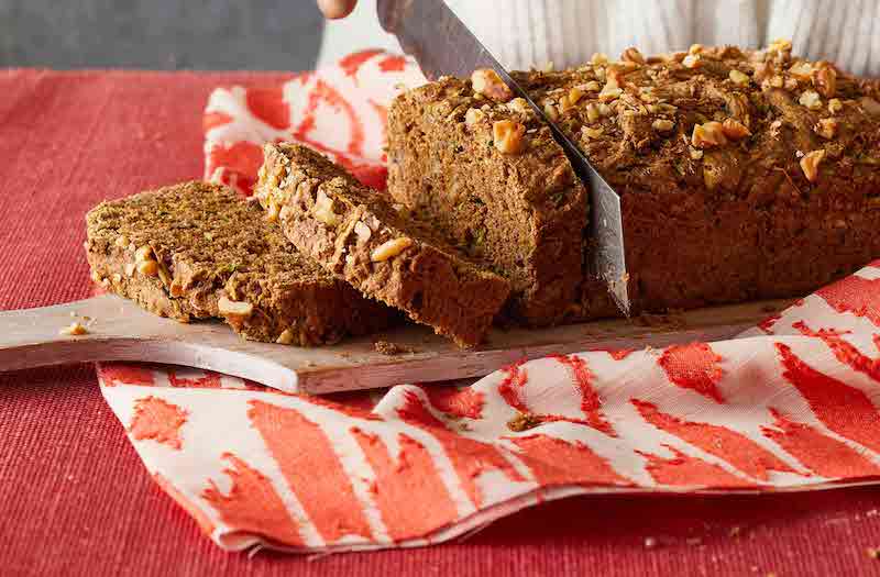 Zucchini Bread being sliced on a cutting board on top of a red cloth on a red table